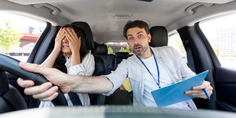 Instructor looking worried and grabbing the steering wheel, with learner covering her eyes